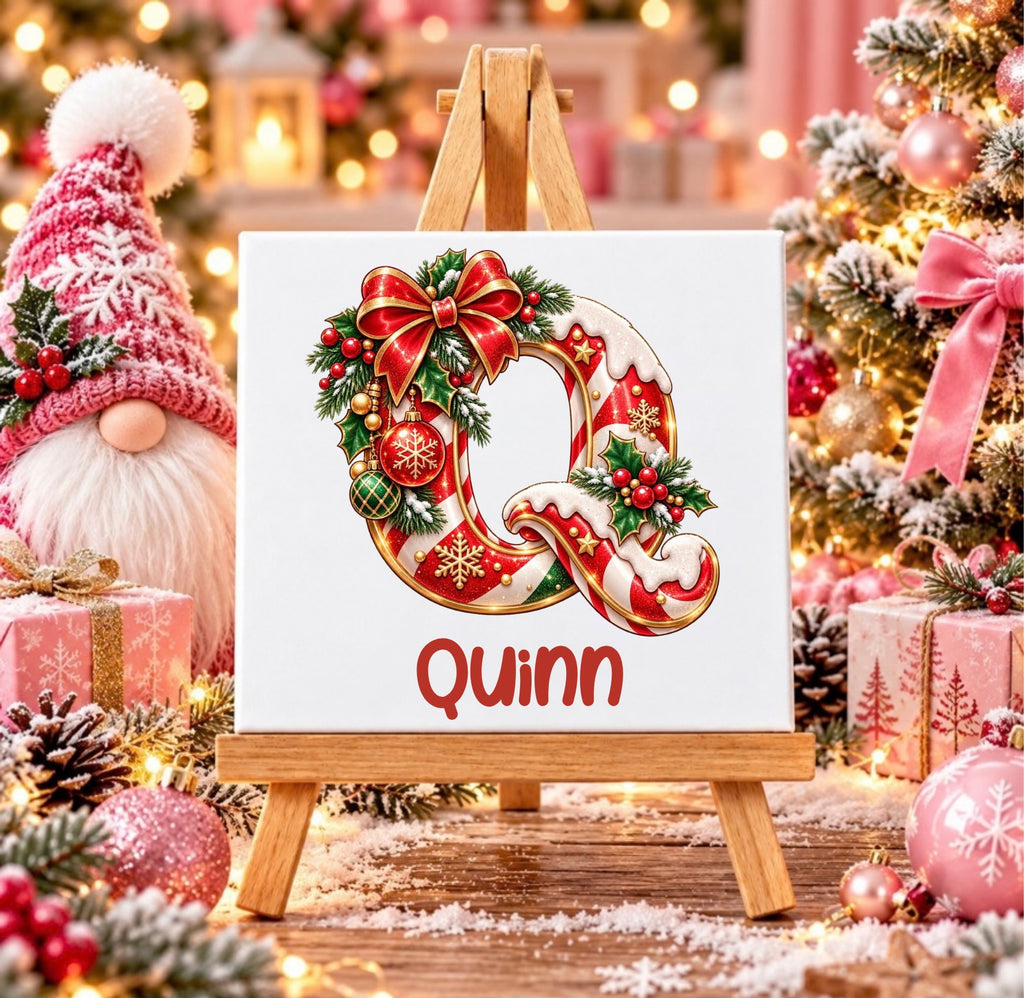 A square tile on a small wooden easel, featuring a festive Christmas letter Q decorated with red bows, green pine branches, and ornaments, with the name 'Quinn' written below in red. The scene is a cozy, decorated Christmas setting with a gnome, wrapped gifts, and a lit Christmas tree in the background.