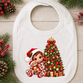A white baby bib with a festive Christmas illustration of a child in a Santa hat hugging a teddy bear next to a decorated Christmas tree, shown on a light wood background with pine branches and berries.