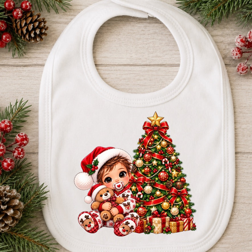 A white baby bib with a festive Christmas illustration of a child in a Santa hat hugging a teddy bear next to a decorated Christmas tree, shown on a light wood background with pine branches and berries.