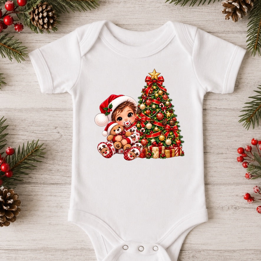 A white short-sleeve baby bodysuit laid flat on a light wood surface, decorated with a colorful Christmas graphic of a child in a Santa hat next to a decorated Christmas tree and gifts. Pine branches, pinecones, and red berries are arranged around the garment.