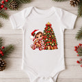 A white short-sleeve baby bodysuit laid flat on a light wood surface, decorated with a colorful Christmas graphic of a child in a Santa hat next to a decorated Christmas tree and gifts. Pine branches, pinecones, and red berries are arranged around the garment.