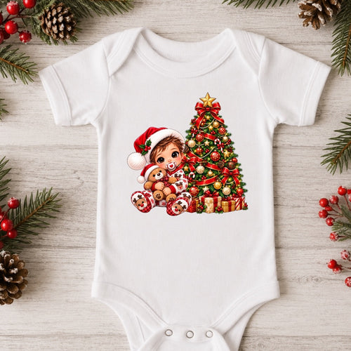 A white short-sleeve baby bodysuit laid flat on a light wood surface, decorated with a colorful Christmas graphic of a child in a Santa hat next to a decorated Christmas tree and gifts. Pine branches, pinecones, and red berries are arranged around the garment.