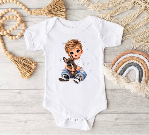 A white short-sleeve baby bodysuit laid flat on a light wood surface, featuring a printed illustration of a blond boy holding a small dog. The bodysuit is styled with boho props including a wooden bead garland, a woven tassel, a faux straw hat, and a small rainbow decoration.
