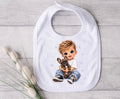 A white baby bib with a custom cartoon illustration of a blond boy hugging a small brown French bulldog, displayed on a light gray wood background next to decorative cotton stems.