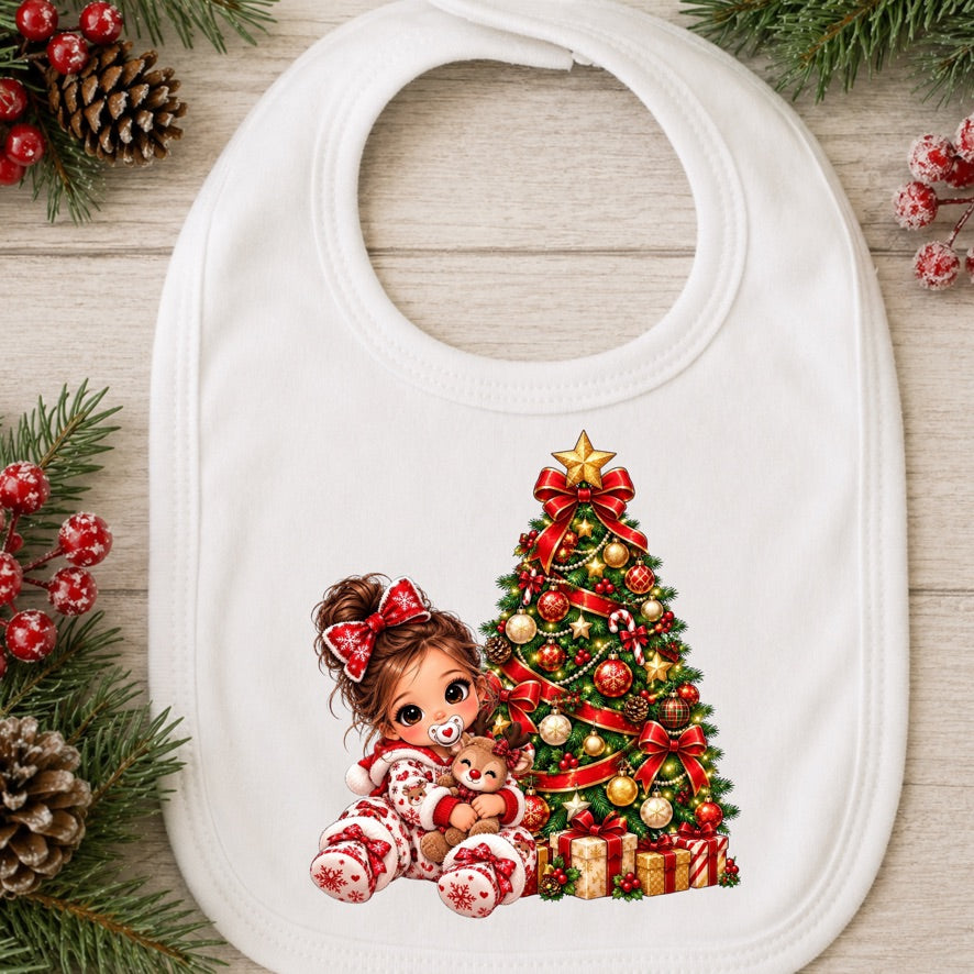 A white baby bib with a festive Christmas graphic, displayed on a light wood background with holiday decorations like pine branches, pinecones, and red berries. The graphic shows a young girl in red and white holiday pajamas sitting next to a decorated Christmas tree with ornaments and gifts.