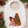 A white baby bib with a festive Christmas graphic, displayed on a light wood background with holiday decorations like pine branches, pinecones, and red berries. The graphic shows a young girl in red and white holiday pajamas sitting next to a decorated Christmas tree with ornaments and gifts.