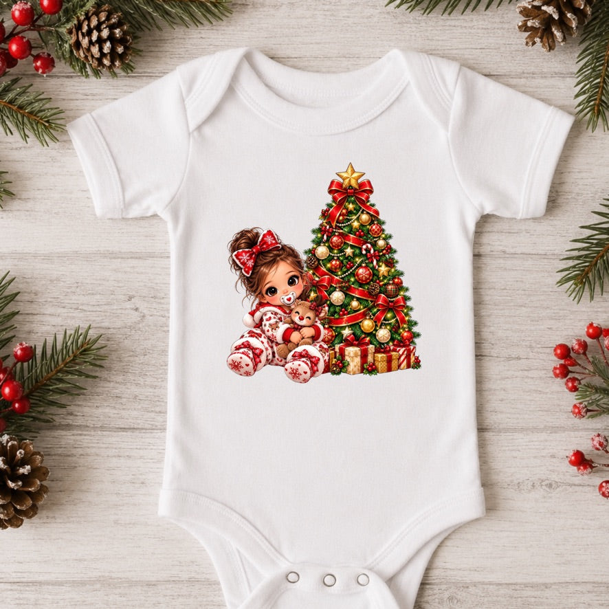 A white short-sleeve baby bodysuit laid flat on a light wood surface, decorated with Christmas greenery, pinecones, and red berries around it. The bodysuit features a colorful graphic of a girl in red-and-white striped pajamas sitting beside a decorated Christmas tree.