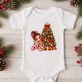 A white short-sleeve baby bodysuit laid flat on a light wood surface, decorated with Christmas greenery, pinecones, and red berries around it. The bodysuit features a colorful graphic of a girl in red-and-white striped pajamas sitting beside a decorated Christmas tree.