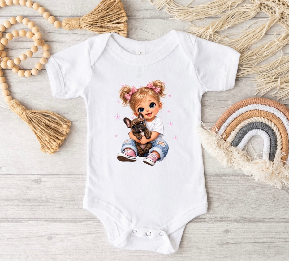A white short-sleeve baby bodysuit laid flat on a light wood surface, featuring a printed illustration of a girl with blonde pigtails holding a small dog. The bodysuit is styled with boho props including a wooden bead garland, a woven tassel, and a small rainbow mat.