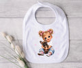 A white baby bib with a printed illustration of a blonde girl with pigtails hugging a small brown puppy, styled on a light gray wood background with decorative bunny tail grass.