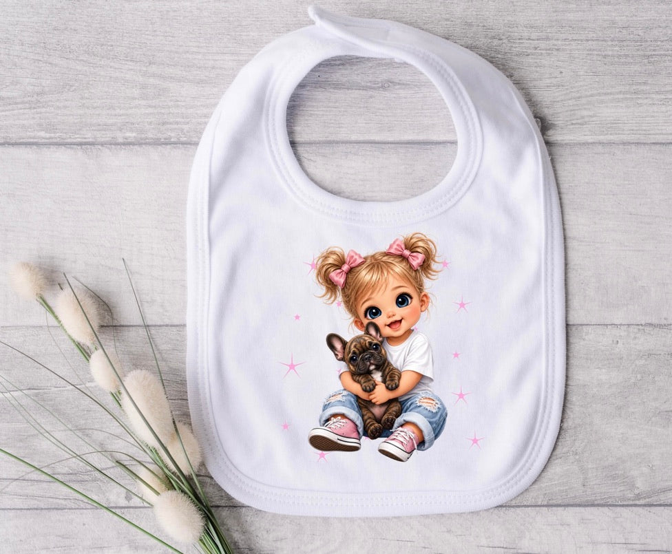 A white baby bib with a printed illustration of a blonde girl with pigtails hugging a small brown puppy, styled on a light gray wood background with decorative bunny tail grass.