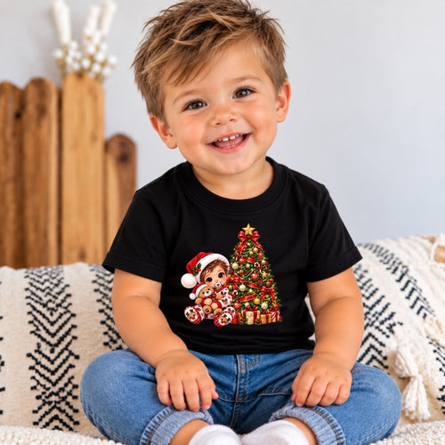 A smiling toddler sitting on a patterned couch wearing a black short-sleeve t-shirt with a Christmas graphic of a baby Jesus and a decorated tree, paired with blue jeans.
