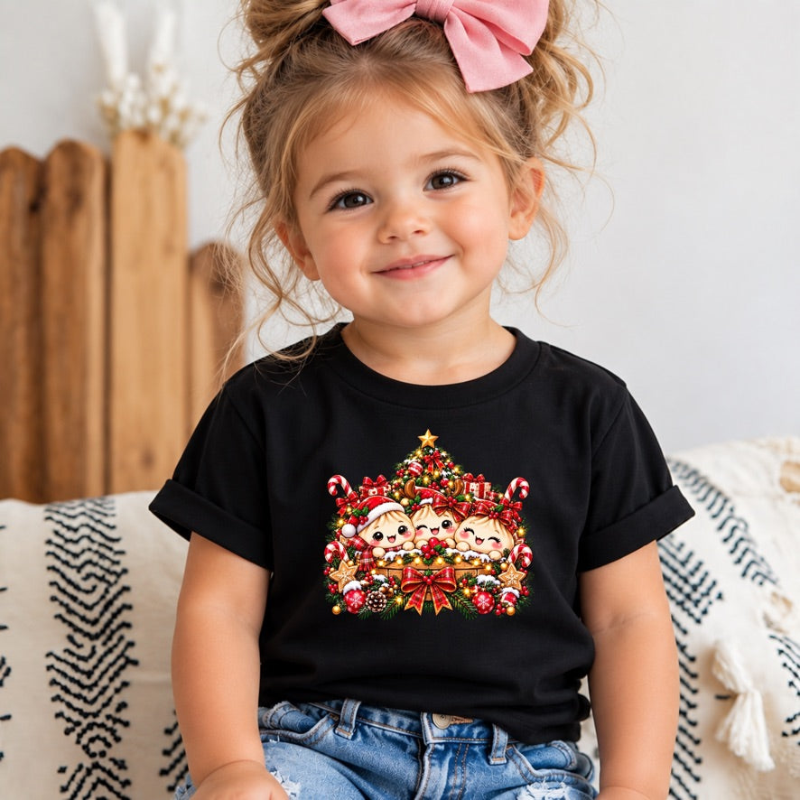 A young child wearing a black short-sleeve t-shirt with a colorful cartoon Christmas tree graphic on the front, paired with blue jeans and a pink hair bow, sitting on a patterned couch.