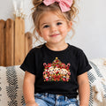 A young child wearing a black short-sleeve t-shirt with a colorful cartoon Christmas tree graphic on the front, paired with blue jeans and a pink hair bow, sitting on a patterned couch.
