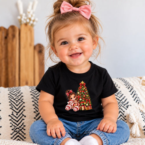 A smiling toddler girl with a pink bow in her hair sits on a patterned throw blanket wearing a black short-sleeve t-shirt with a Christmas tree and doll graphic, paired with blue jeans.