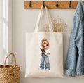 A cream canvas tote bag hanging on a wooden wall hook, featuring a cartoon illustration of a girl with blonde hair holding a small black dog, wearing blue jeans and a white t-shirt with pink sparkles. A denim jacket hangs on the right, and a woven basket is on the left.