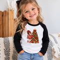 A young girl with blonde hair in a ponytail smiles while wearing a black and white raglan baseball t-shirt with a Christmas-themed graphic of a girl and a decorated tree. She is sitting on a patterned blanket in a cozy indoor setting.
