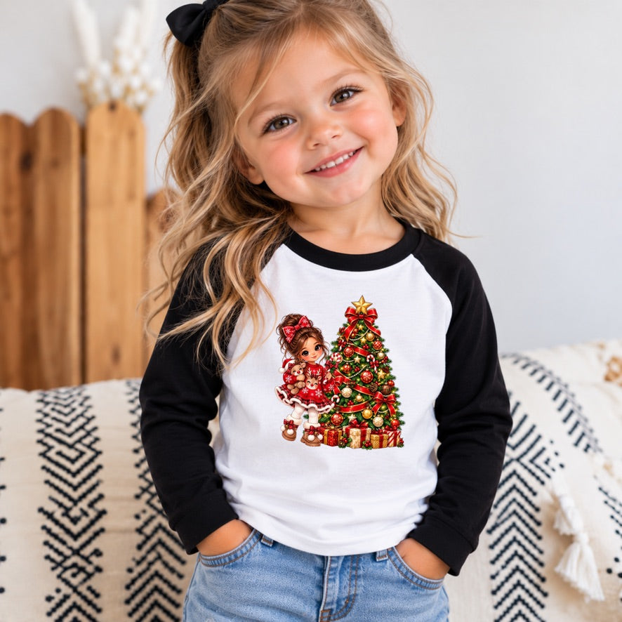 A young girl with blonde hair in a ponytail smiles while wearing a black and white raglan baseball t-shirt with a Christmas-themed graphic of a girl and a decorated tree. She is sitting on a patterned blanket in a cozy indoor setting.