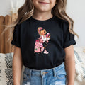 A young girl wearing a black short-sleeve t-shirt with a colorful cartoon graphic of a girl with pink luggage and a drink, paired with blue jeans.