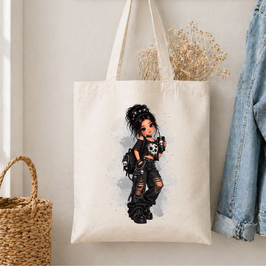 A cream canvas tote bag hanging on a wall, featuring a printed illustration of a gothic girl with black hair, a skull necklace, and ripped black clothes. The bag is styled with dried flowers, a woven basket, and a denim jacket in the background.