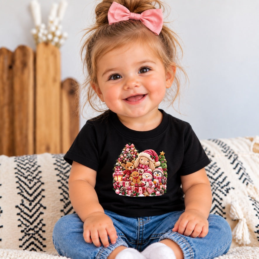 A smiling toddler girl with a pink bow in her hair sits on a patterned throw blanket wearing a black short-sleeve t-shirt with a colorful Christmas-themed graphic print and blue jeans.