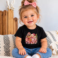 A smiling toddler girl with a pink bow in her hair sits on a patterned throw blanket wearing a black short-sleeve t-shirt with a colorful Christmas-themed graphic print and blue jeans.