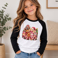 A young girl with long wavy brown hair smiling, wearing a white and black raglan long-sleeve shirt with a colorful Christmas teddy bear graphic on the front, paired with blue jeans in a bright indoor setting.