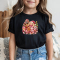 A young girl wearing a black short-sleeve t-shirt with a colorful Christmas-themed cartoon graphic on the chest, paired with light blue jeans.