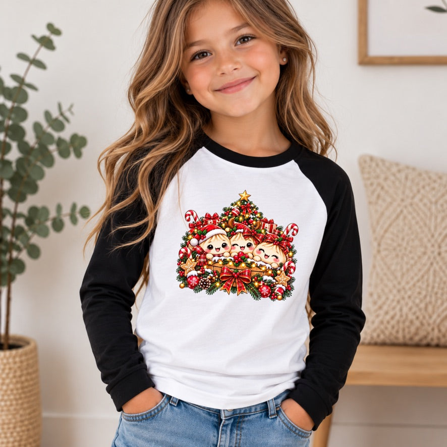 A young girl with long wavy brown hair smiling, wearing a black and white raglan long-sleeve shirt with a colorful Christmas tree graphic on the front, paired with blue jeans in a bright indoor setting.