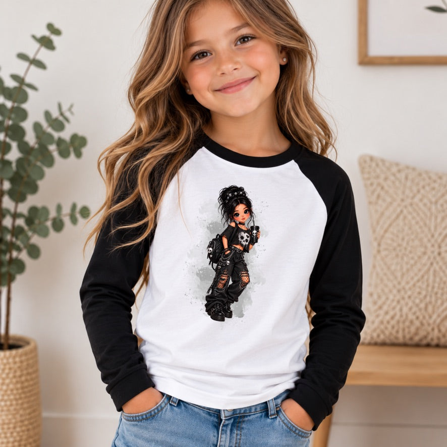 A young girl with long wavy brown hair smiling while wearing a black and white raglan baseball t-shirt with a graphic of a doll on the front, paired with blue jeans in a brightly lit indoor setting.