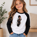 A young girl with long wavy brown hair smiling while wearing a black and white raglan baseball t-shirt with a graphic of a doll on the front, paired with blue jeans in a brightly lit indoor setting.