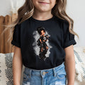 A young girl wearing a black short-sleeve t-shirt with a graphic of a punk-style cartoon girl, paired with light blue jeans, shown in a cozy indoor setting.