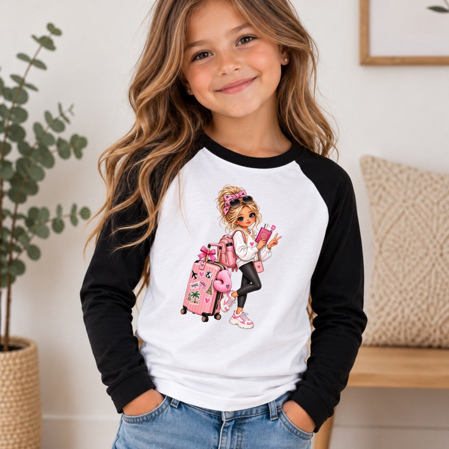 A young girl with long wavy brown hair smiling, wearing a black and white raglan long-sleeve shirt with a colorful cartoon graphic of a girl with pink luggage on the front, paired with blue jeans in a brightly lit indoor setting.