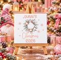 Personalised first Christmas ceramic tile with name and year, featuring a blush pink festive wreath and snowflakes displayed on a wooden stand.
