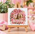 A square white canvas print on a small wooden easel, featuring a pink and rose 21st birthday design with a photo of a young woman, set on a wooden table with pink decorative props.