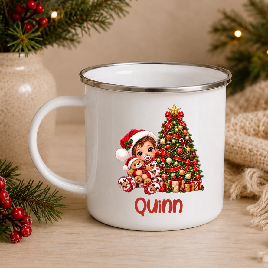 A white enamel-style mug with a silver rim, decorated with a Christmas tree, teddy bear, Santa hat, and the name 'Quinn' in red. The mug sits on a wooden surface surrounded by holiday greenery, red berries, and a beige burlap fabric.