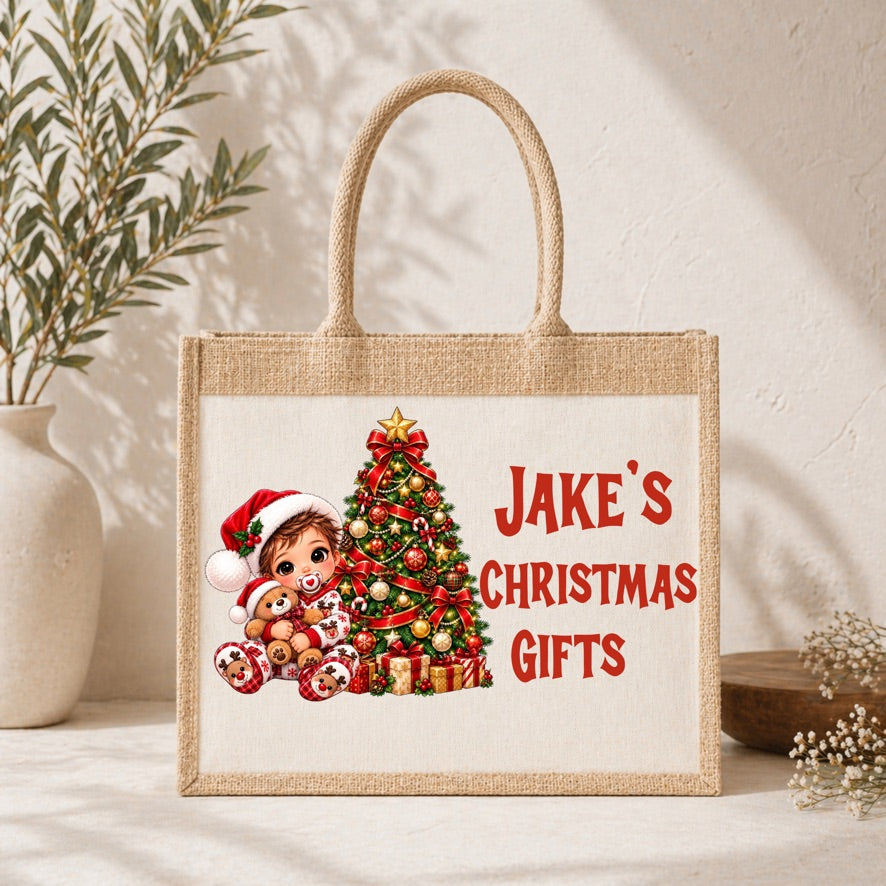 A natural jute tote bag with rope handles, featuring a personalized Christmas illustration of a child, teddy bear, and a decorated tree next to the red text 'JAKE'S CHRISTMAS GIFTS'. The bag is styled on a white surface with a textured wall background, a potted plant, and small flowers.
