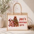 A natural jute tote bag with rope handles, featuring a personalized Christmas illustration of a child, teddy bear, and a decorated tree next to the red text 'JAKE'S CHRISTMAS GIFTS'. The bag is styled on a white surface with a textured wall background, a potted plant, and small flowers.