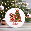 A personalized round white Christmas ornament featuring an illustration of a child in a Santa hat next to a decorated Christmas tree, with the name 'Jake' printed in red at the bottom. The ornament hangs from a gold string against a blurred festive background with a Christmas tree and lights.