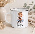 A white enamel-style mug with a silver rim, featuring a printed illustration of a boy holding a small dog and the name 'Jake' below. The mug is styled on a light wood surface with dried flowers, a ceramic vase, and a knitted textile in the background.