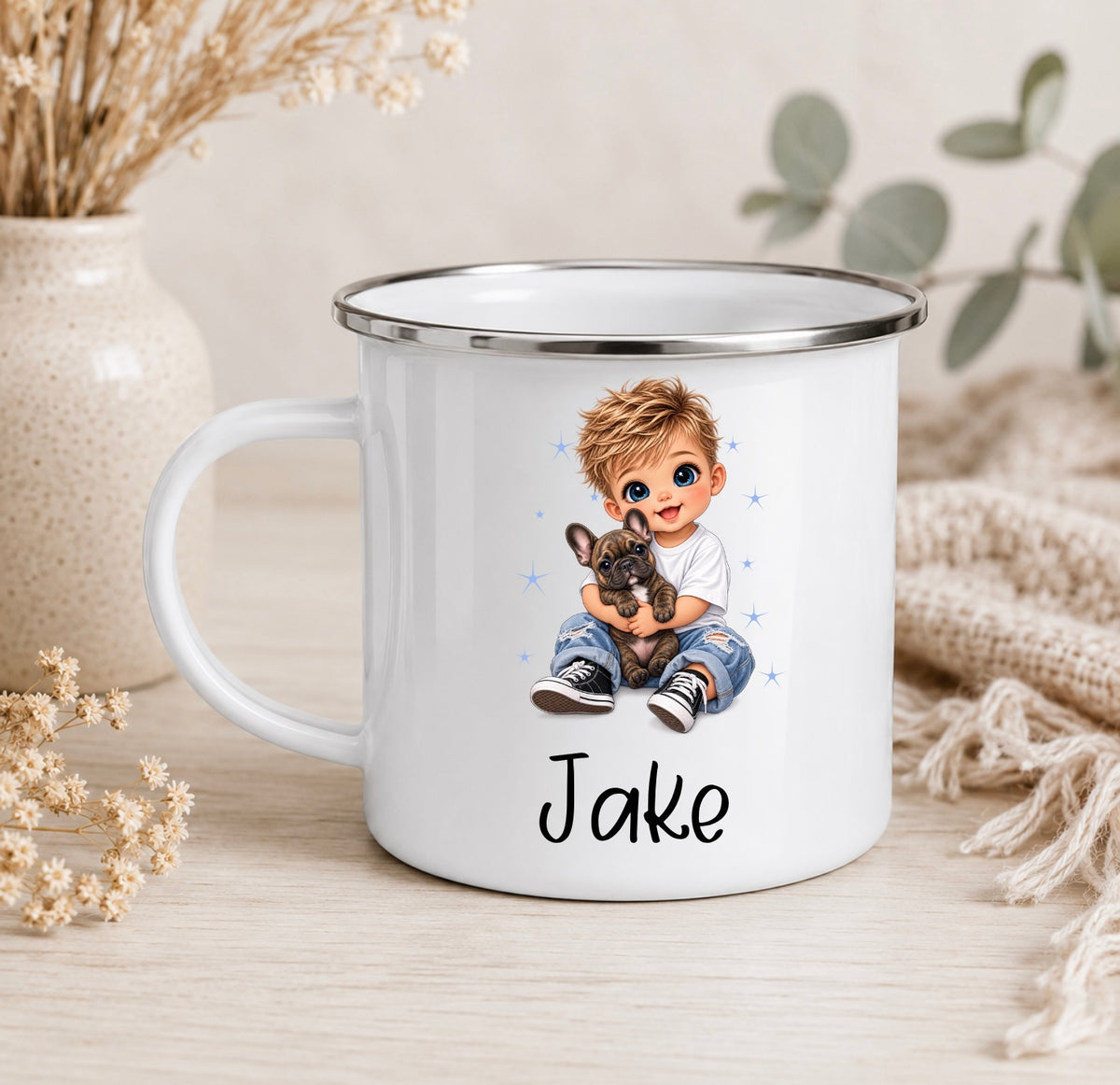 A white enamel-style mug with a silver rim, featuring a printed illustration of a boy holding a small dog and the name 'Jake' below. The mug is styled on a light wood surface with dried flowers, a ceramic vase, and a knitted textile in the background.