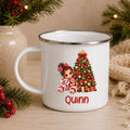 A white enamel-style mug with a silver rim, decorated with a Christmas tree illustration, a doll, and the name 'Quinn' in red. The mug is styled on a wooden surface with holiday greenery, red berries, and a burlap fabric in the background.