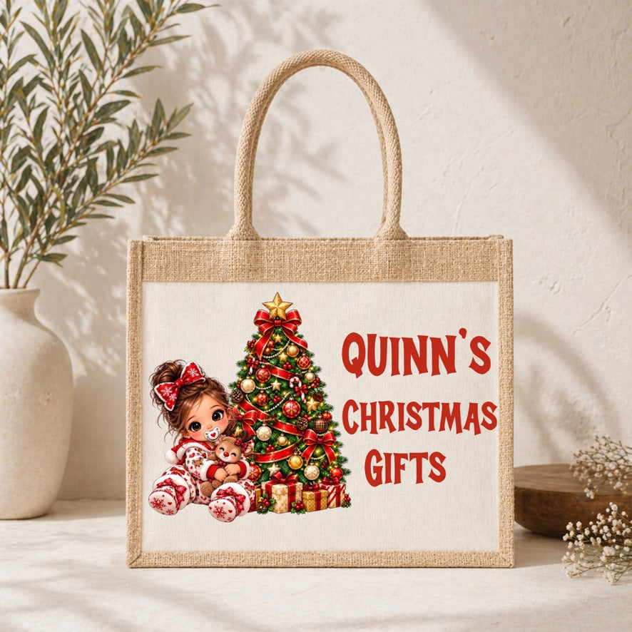 A natural jute tote bag with a printed Christmas illustration and the text 'QUINN'S CHRISTMAS GIFTS' in red, styled on a light surface with decorative plants in the background.