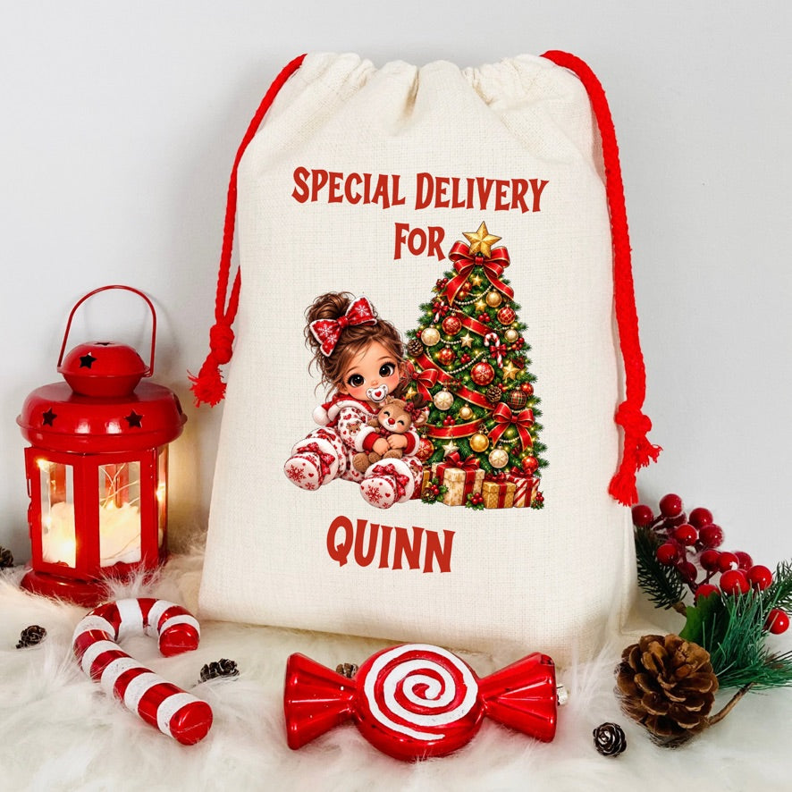 A personalized cream canvas Christmas gift sack with red drawstrings, featuring a printed illustration of a girl and a decorated Christmas tree with the text 'SPECIAL DELIVERY FOR QUINN'. The sack is styled on a white faux fur surface with red holiday props including a lantern, candy canes, a lollipop-shaped ornament, pinecones, and a berry sprig.