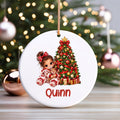 A round white Christmas ornament with a hanging loop, featuring an illustration of a young girl in pajamas sitting by a decorated Christmas tree and the name 'Quinn' in red text at the bottom. The ornament is displayed on a wooden surface with a blurred Christmas tree and ornaments in the background.