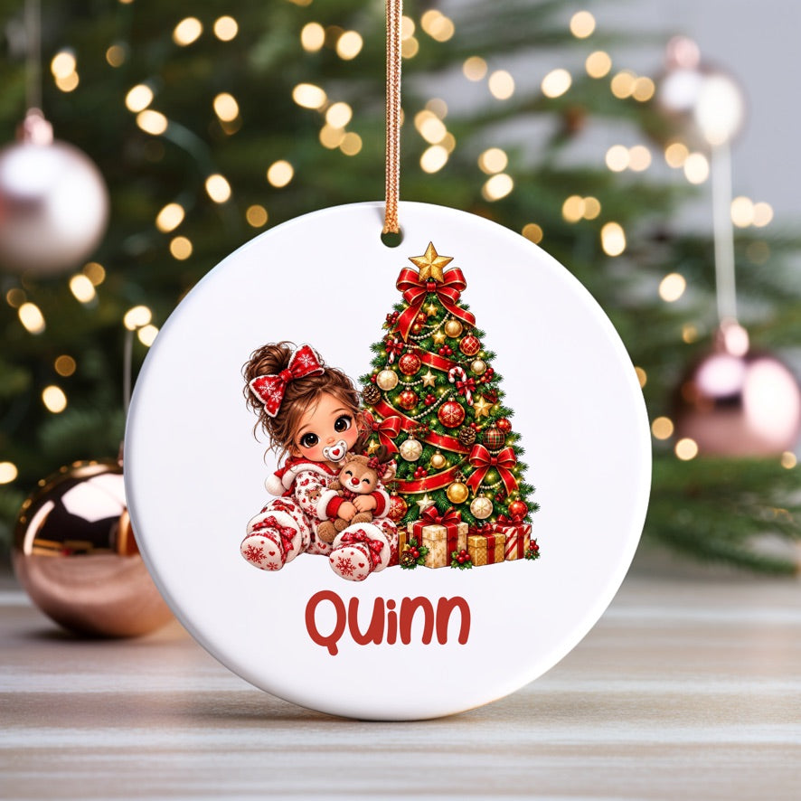 A round white Christmas ornament with a hanging loop, featuring an illustration of a young girl in pajamas sitting by a decorated Christmas tree and the name 'Quinn' in red text at the bottom. The ornament is displayed on a wooden surface with a blurred Christmas tree and ornaments in the background.