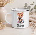 A white enamel-style mug with a silver rim, featuring a printed illustration of a girl with pigtails holding a dog and the name 'Quinn' below. The mug is styled on a light wood surface with dried flowers, a vase, and a knitted textile in the background.