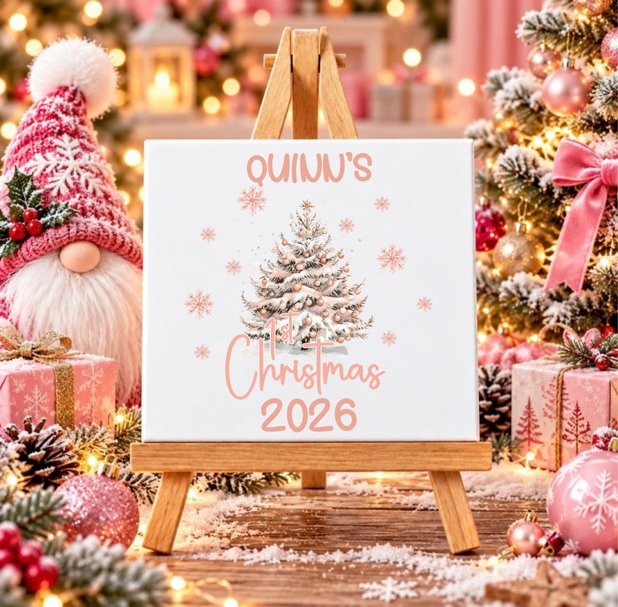 A personalized white canvas print with a Christmas tree illustration and the text 'QUINN'S Christmas 2026' displayed on a small wooden easel. The scene is decorated for Christmas with a gnome, ornaments, wrapped gifts, and a lit tree in the background.