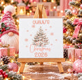 A personalized white canvas print with a Christmas tree illustration and the text 'QUINN'S Christmas 2026' displayed on a small wooden easel. The scene is decorated for Christmas with a gnome, ornaments, wrapped gifts, and a lit tree in the background.