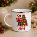 A white enamel-style mug with a silver rim, featuring a printed illustration of a child in a Santa hat next to a decorated Christmas tree and the name 'Jake' in red. The mug is styled on a wooden surface with holiday greenery and burlap in the background.